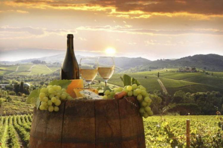 Foto panoramica al tramonto di vigneti e colline con una botte in primo piano su cui sono posati 2 calici di vino e bottiglia tra ciocche di uva
